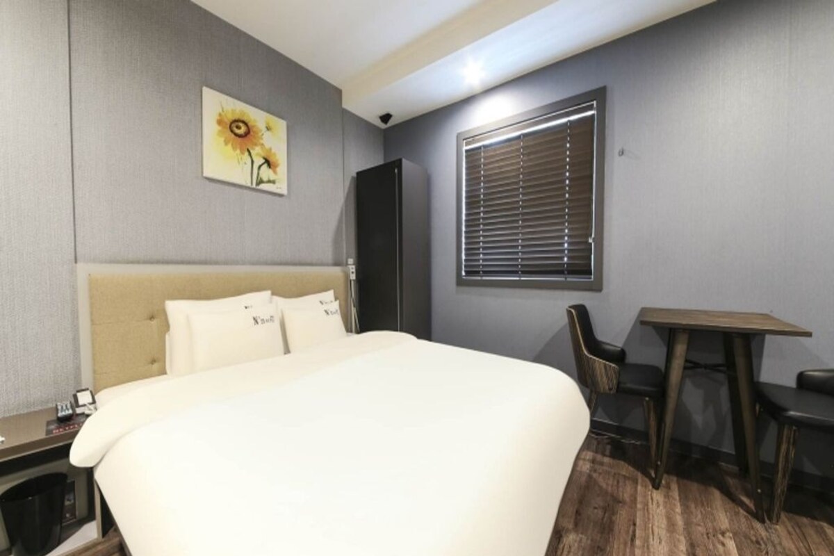 A hotel room with a bed, a desk, a chair, a window with blinds, and a painting on the wall.