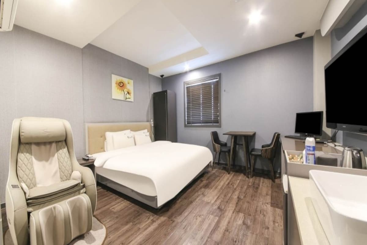A hotel room with a bed, a massage chair, a small table, a TV, and a bathroom with a sink and mirror.