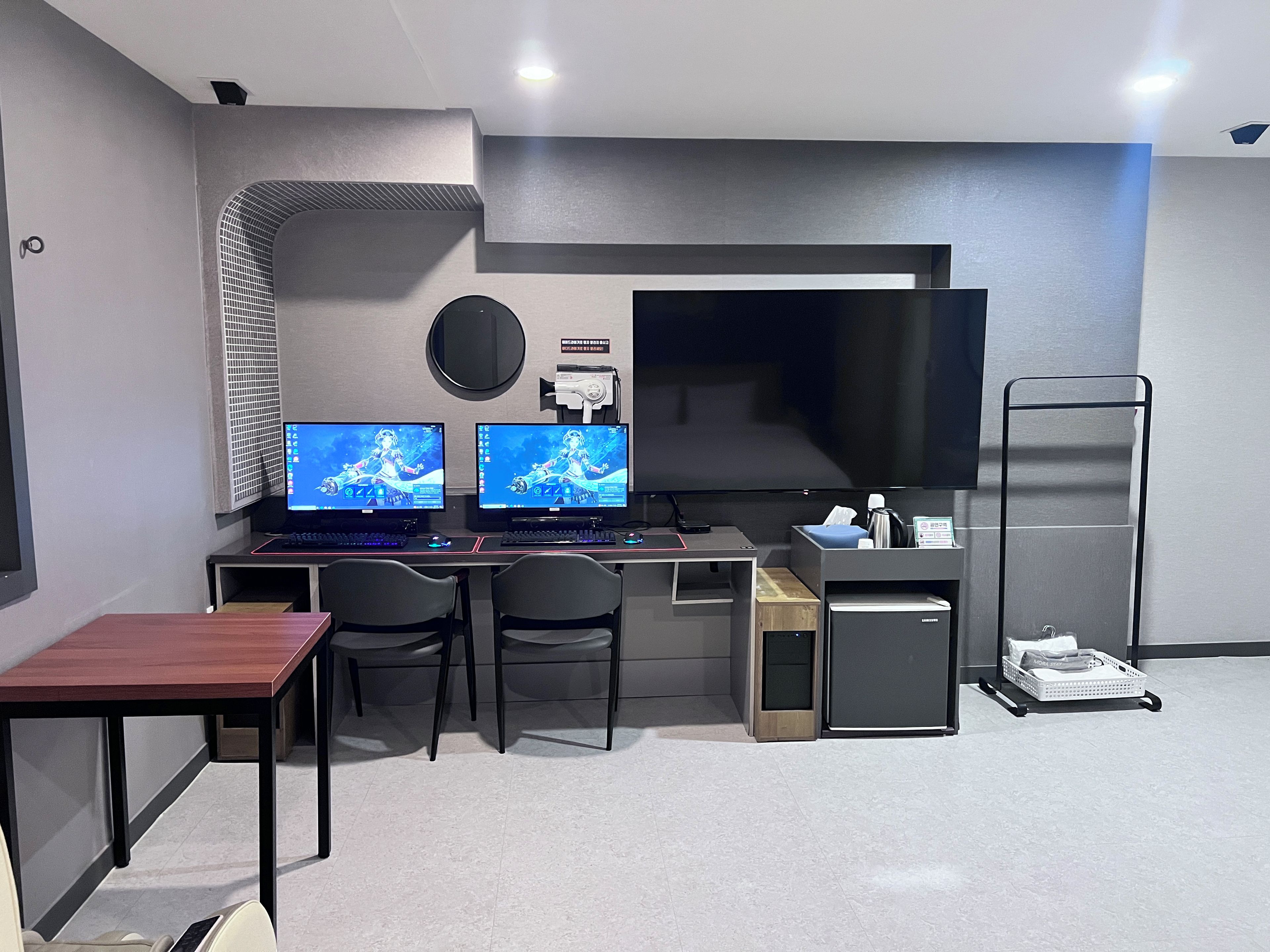 Una habitación moderna con dos monitores, un escritorio, sillas, un televisor de pantalla plana, un refrigerador pequeño y un carrito con objetos.