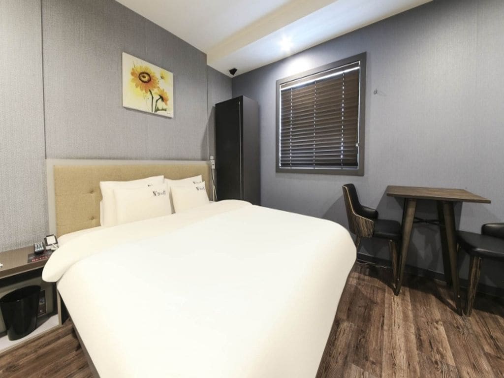 A hotel room with a bed, a desk, a chair, a window with blinds, and a painting on the wall.