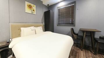 A hotel room with a bed, a desk, a chair, a window with blinds, and a painting on the wall.