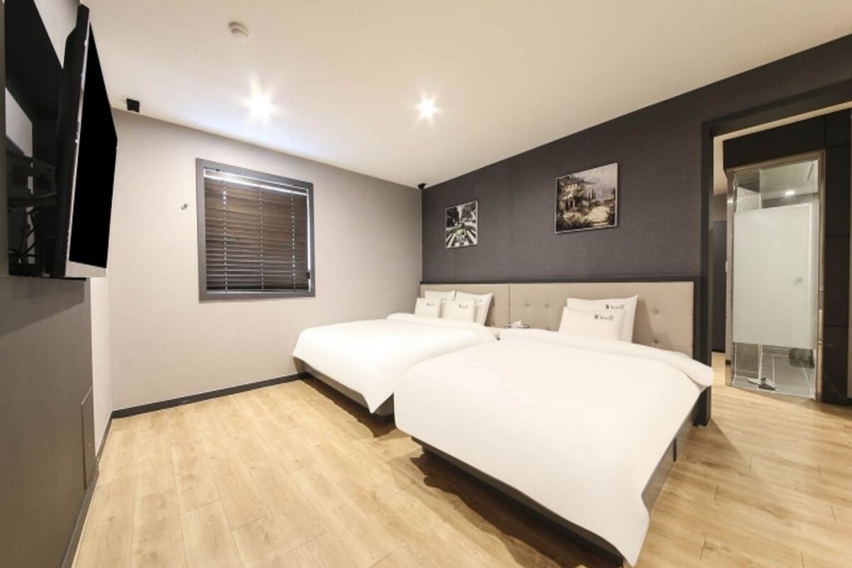 A bedroom with two beds, a window with blinds, and a flat-screen TV mounted on the wall.