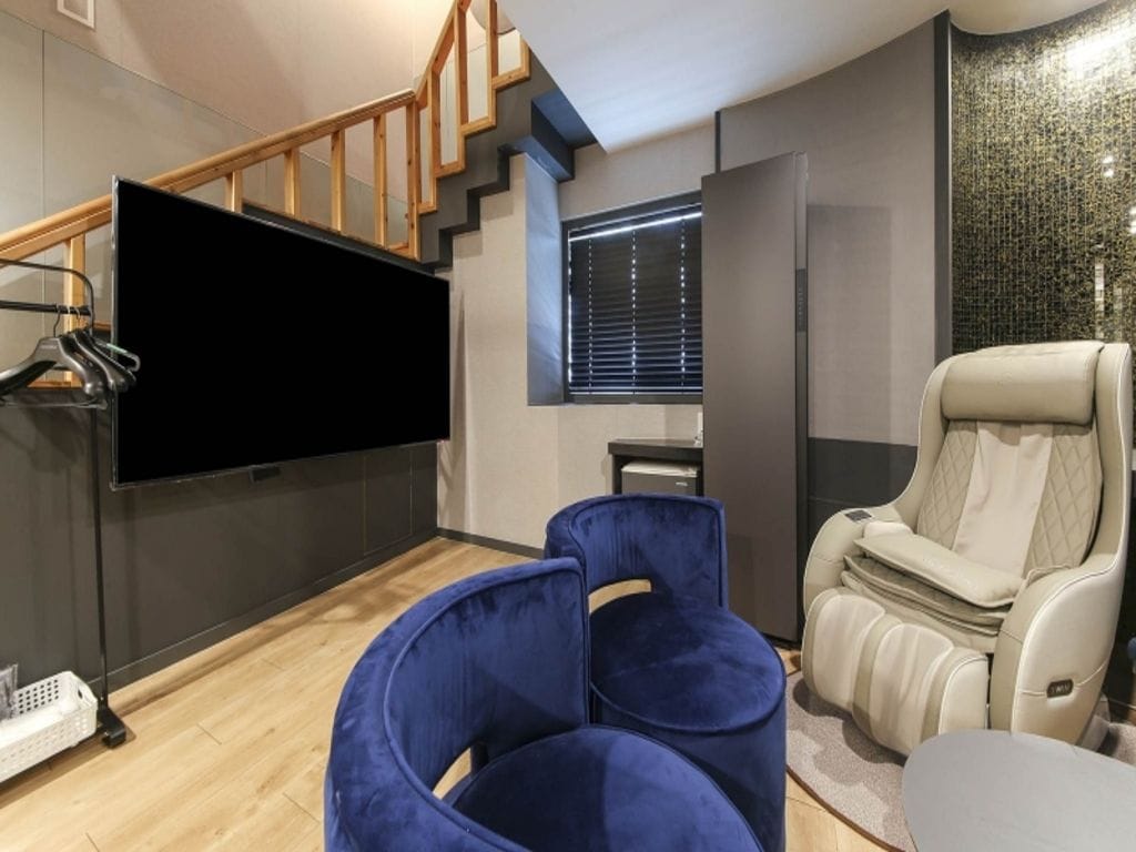A modern living room with a flat-screen TV, a blue chair, a massage chair, and a staircase.