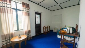 A room with a blue carpet, a wooden table with a bowl, a wooden chair, a bed with a blue cover, a wooden nightstand, and a window with curtains. - The Crown's Lodge Misty Hideaway  (Nuwara eliya)