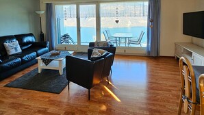 Een woonkamer met een zwarte leren bank, een bijzettafeltje, een zwarte fauteuil en een eethoek met een tafel en stoelen.