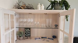 Een houten kast met glazen planken, gevuld met wijnglazen, een krijtbord en decoratieve objecten.