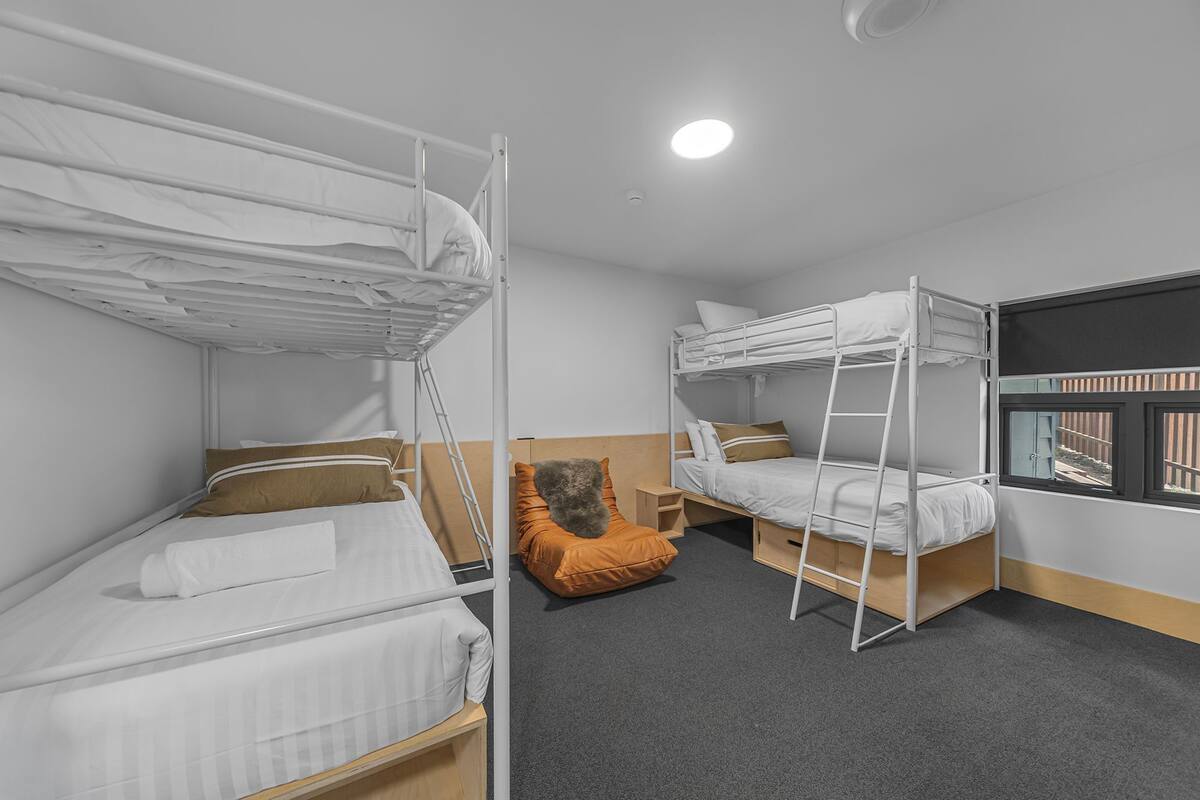 Une chambre avec deux lits superposés, un petit pouf orange, une fenêtre avec vue et un luminaire au plafond.
