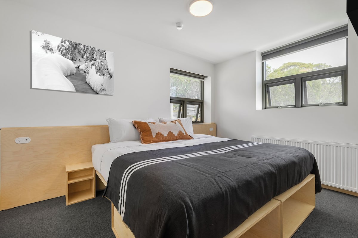 Une chambre à coucher moderne avec une tête de lit en bois, une couvre-lit noir et blanc et une grande fenêtre laissant entrer la lumière naturelle.