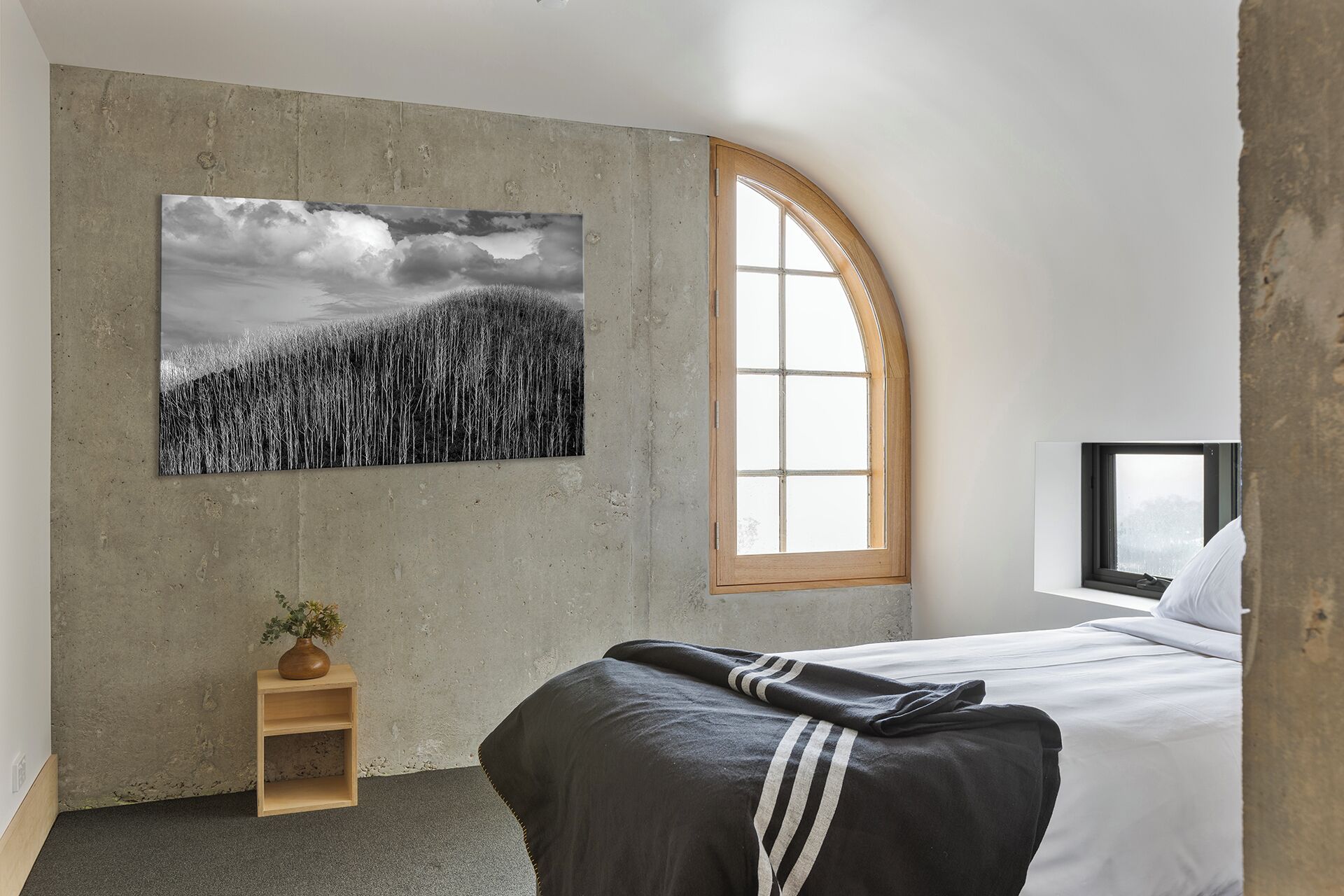 A bedroom with a large window, a bed, a small wooden nightstand, and a painting on the wall.