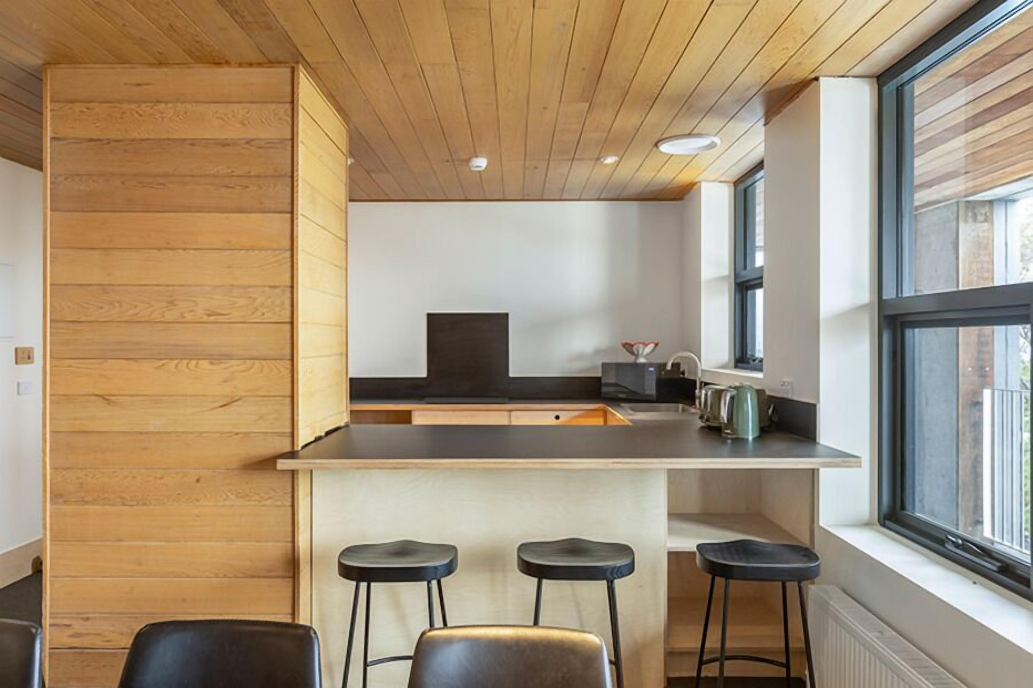 A modern kitchen with wooden paneling, a central island with stools, and large windows.