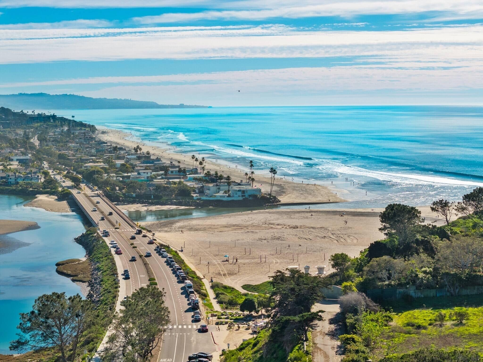 A coastal road with a beach and ocean on the right, a bridge on the left, and a hilly landscape in the background.