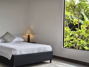A bedroom with a bed, a bedside lamp, and a view of greenery through a window.