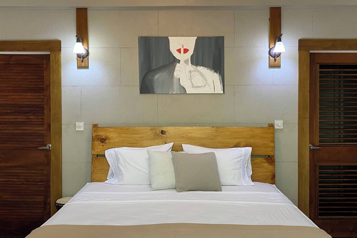 A hotel room with a wooden headboard, a bed with white bedding, a painting on the wall, and two wall-mounted lamps.