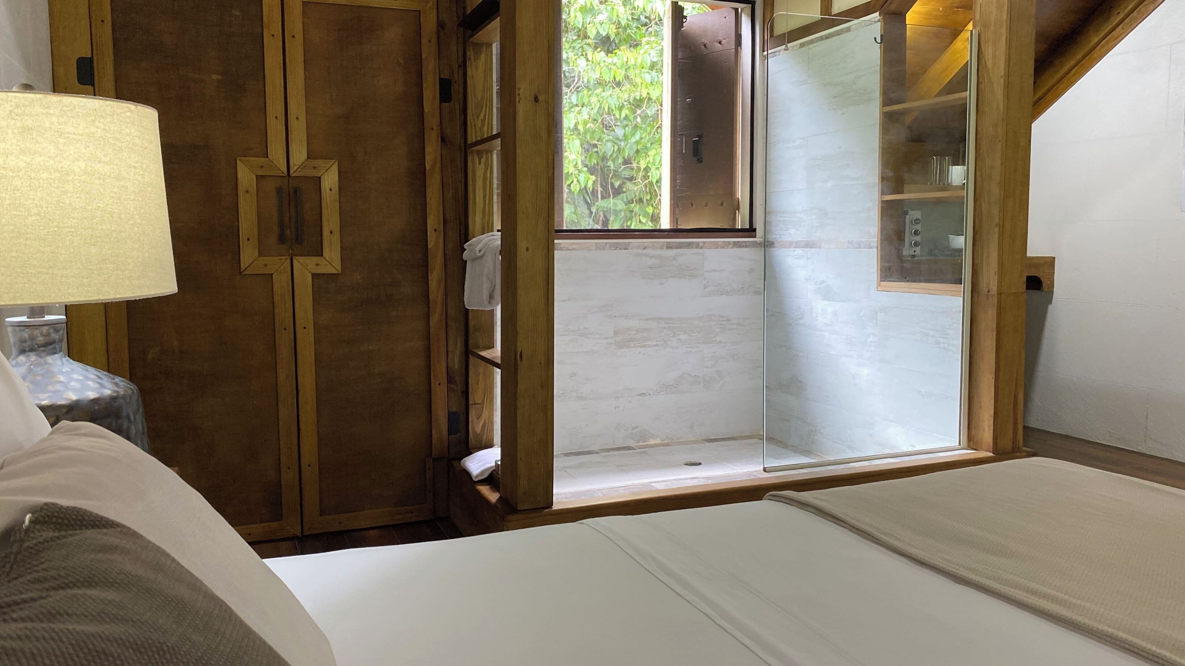 A bedroom with a bed, a nightstand with a lamp, a wooden wardrobe, and a glass shower enclosure.
