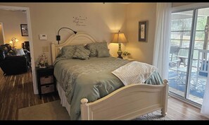 A bedroom with a bed, bedside lamps, a nightstand, a chair, and a view of the outdoors through a sliding glass door.