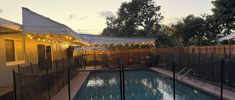 A swimming pool with a fenced deck, a covered patio area with string lights, and a wooden fence in the background.