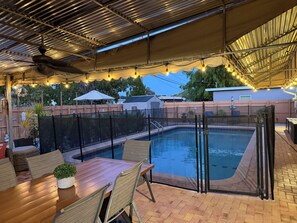 Uma área de piscina ao ar livre com mesa e cadeiras de madeira, cercada por uma grade e coberta por um toldo.