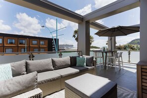 A balcony with a wicker sofa, a table, and chairs, overlooking a cruise ship and a building with 'QUEST' signage. - Elevated Mount Location, Beach, Shops, 2 Car Parks (Mount Maunganui)