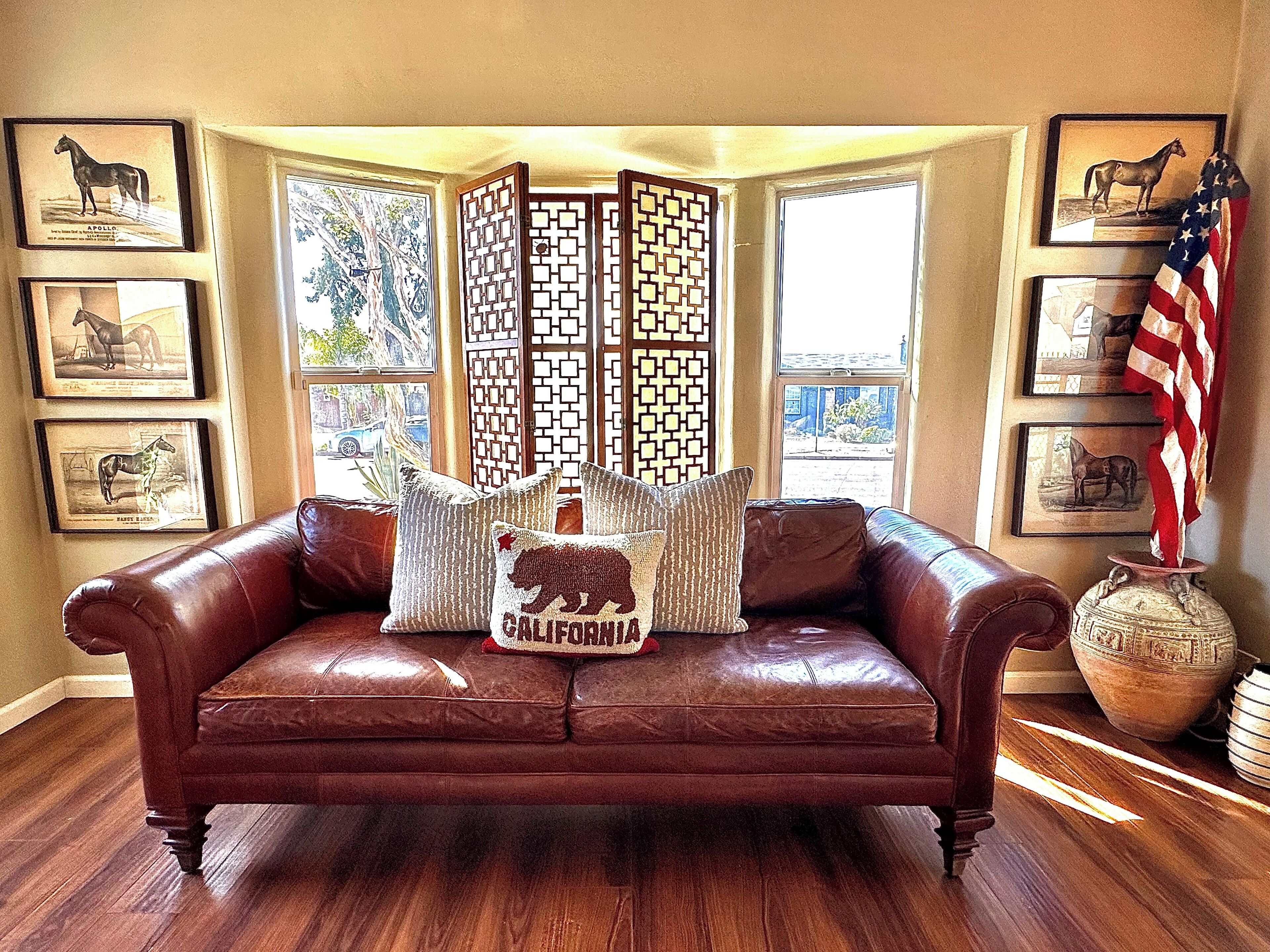 Un canapé en cuir marron agrémenté d’un coussin orné d’un ours de Californie, entouré de portraits équestres encadrés, d’un paravent décoratif et d’un drapeau américain.