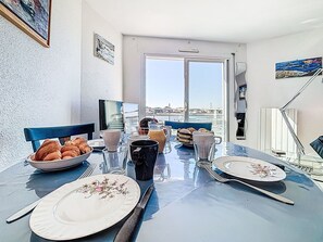 Ein Esstisch mit Tellern, Besteck und einer Kaffeekanne vor einer weißen Wand mit einem Fenster mit Blick auf einen Hafen.