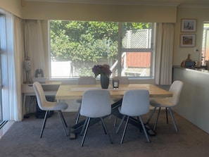 A dining area with a wooden table, six chairs, a vase with flowers, and a view of greenery outside.