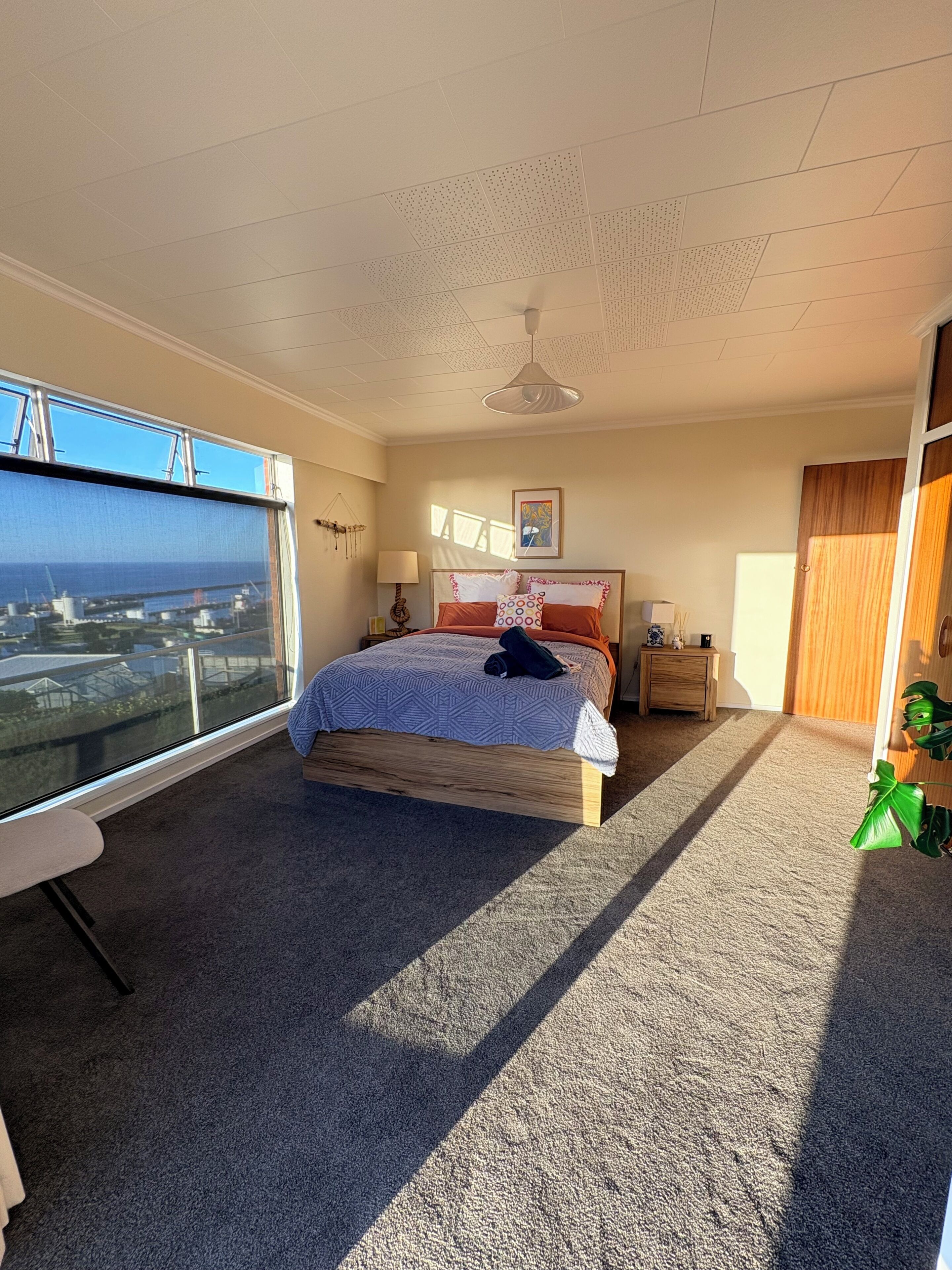 Une chambre avec un grand lit, un ventilateur de plafond, une fenêtre donnant sur un chantier de construction et une porte en bois.
