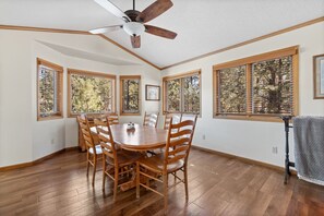 Une salle à manger avec une table et des chaises en bois, de grandes fenêtres équipées de stores et un ventilateur de plafond.