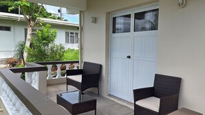 Un balcón con mobiliario de mimbre, una puerta blanca y vista a un edificio con tejado azul.