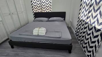 Cama bem arrumada com roupa de cama cinza e uma toalha dobrada por cima, cabeceira preta e parede estampada à direita.