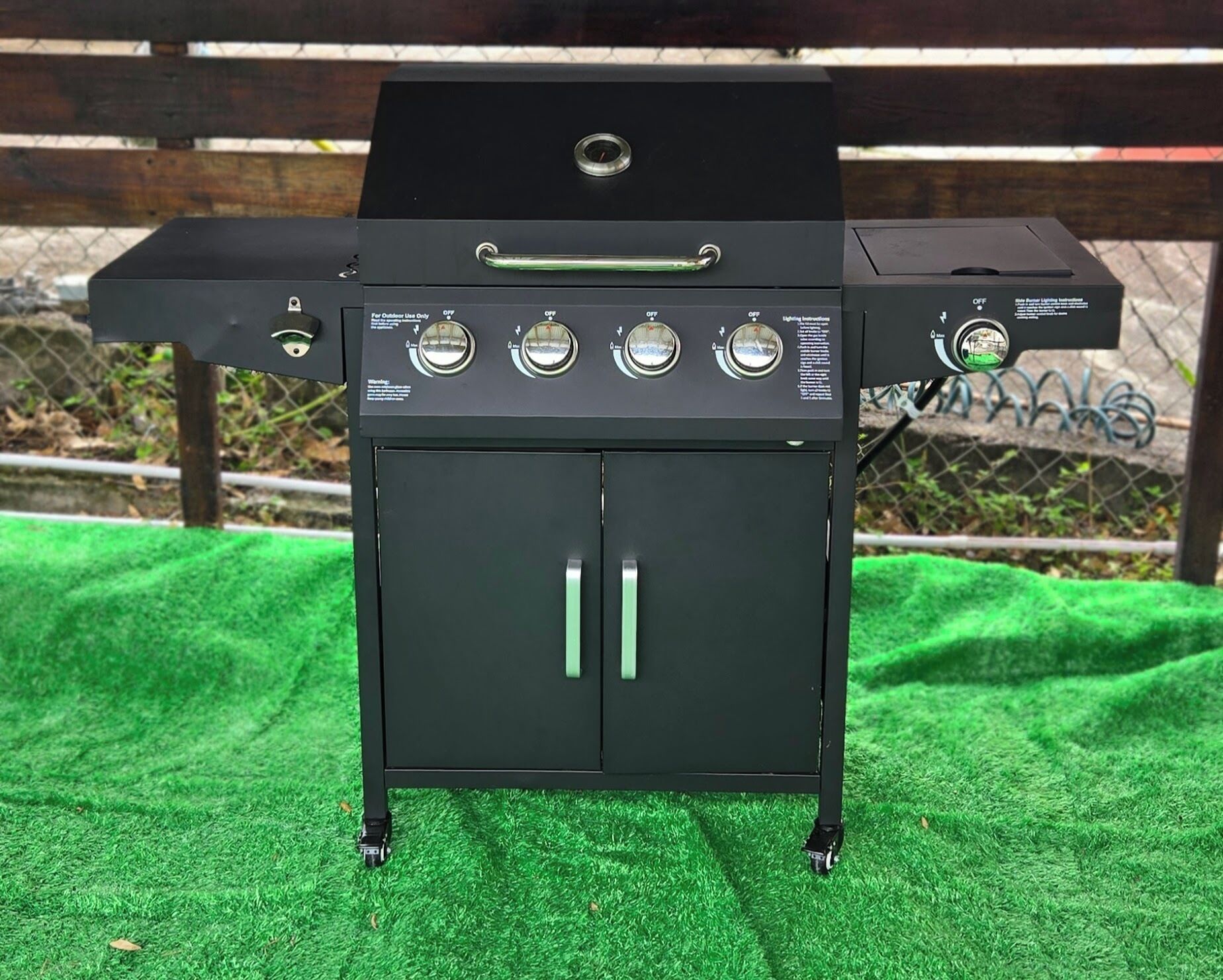 A portable barbecue grill with four control knobs, a handle, and a side burner, set on a green surface.