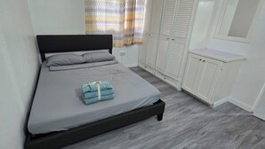 Una cama bien hecha con colchón y almohadas grises, una toalla doblada y un cabecero negro. A la derecha hay un armario blanco y una ventana con cortinas estampadas.