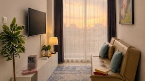 A modern living room with a sofa, a flat-screen TV, a potted plant, a lamp, and a view of the city through large windows.