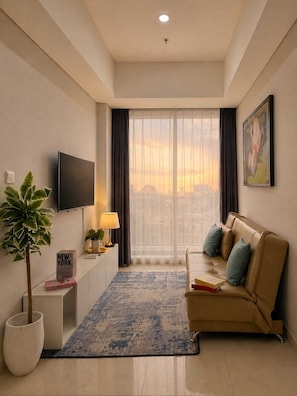 A modern living room with a sofa, a flat-screen TV, a potted plant, a lamp, and a view of the city through large windows. - Teras By SanLiving - 3BR - Hublife Mall (Jakarta)