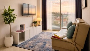 A modern living room with a flat-screen TV, a potted plant, a sofa with cushions, a small side table, and a view of the city through large windows.