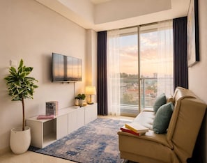 A modern living room with a flat-screen TV, a potted plant, a sofa with cushions, a small side table, and a view of the city through large windows. - Teras By SanLiving - 3BR - Hublife Mall (Jakarta)