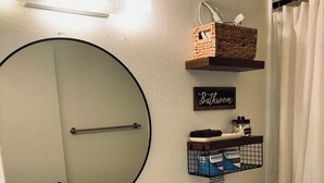 Un baño con un espejo redondo, un lavamanos, un inodoro y una cortina de ducha. Hay repisas de almacenamiento sobre el inodoro y una pequeña planta en maceta sobre el lavamanos.
