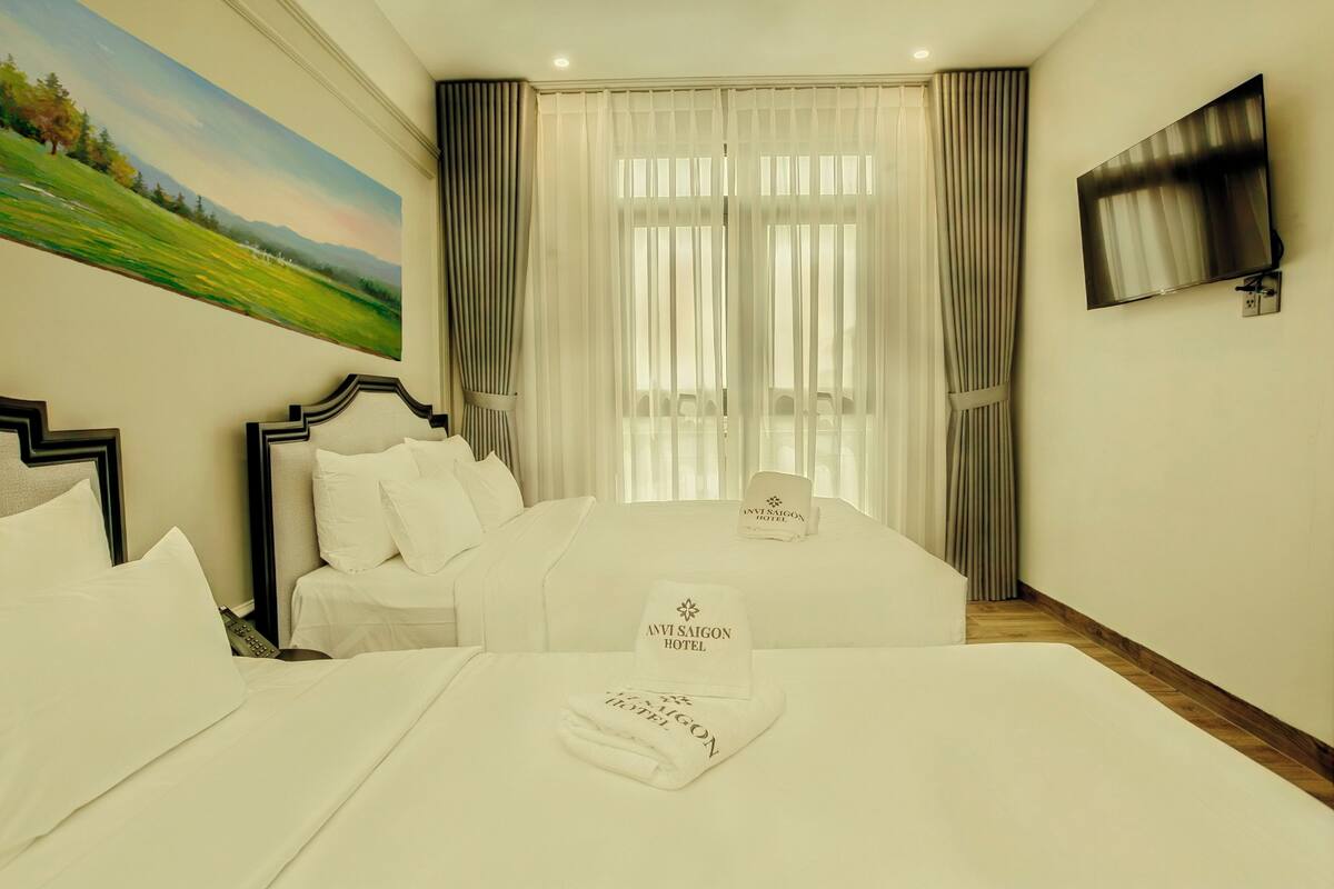 A hotel room with two beds, a painting on the wall, a TV mounted on the wall, and a window with curtains.