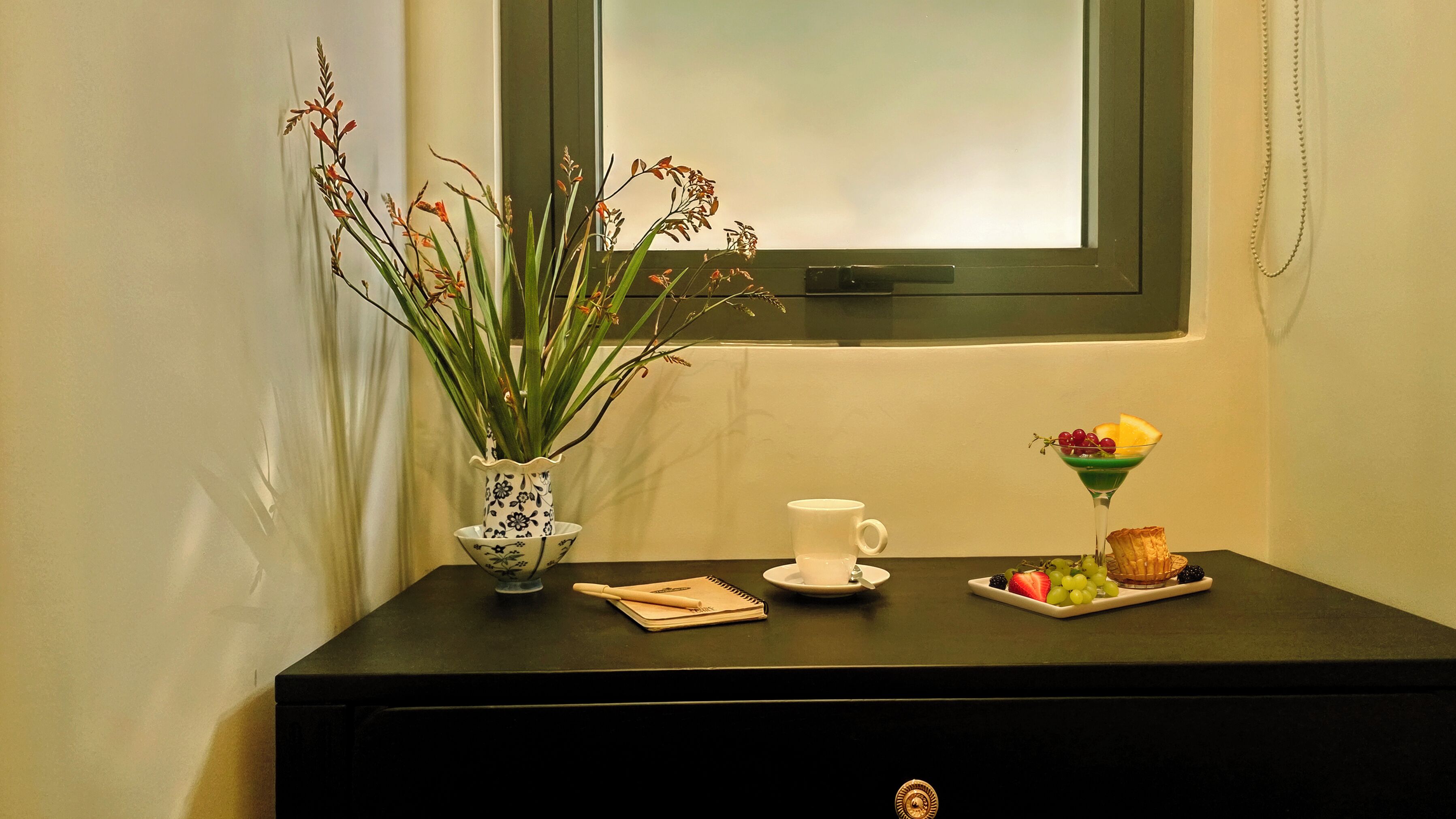 A table with a vase of plants, a cup, a plate of fruit, and a small cake.