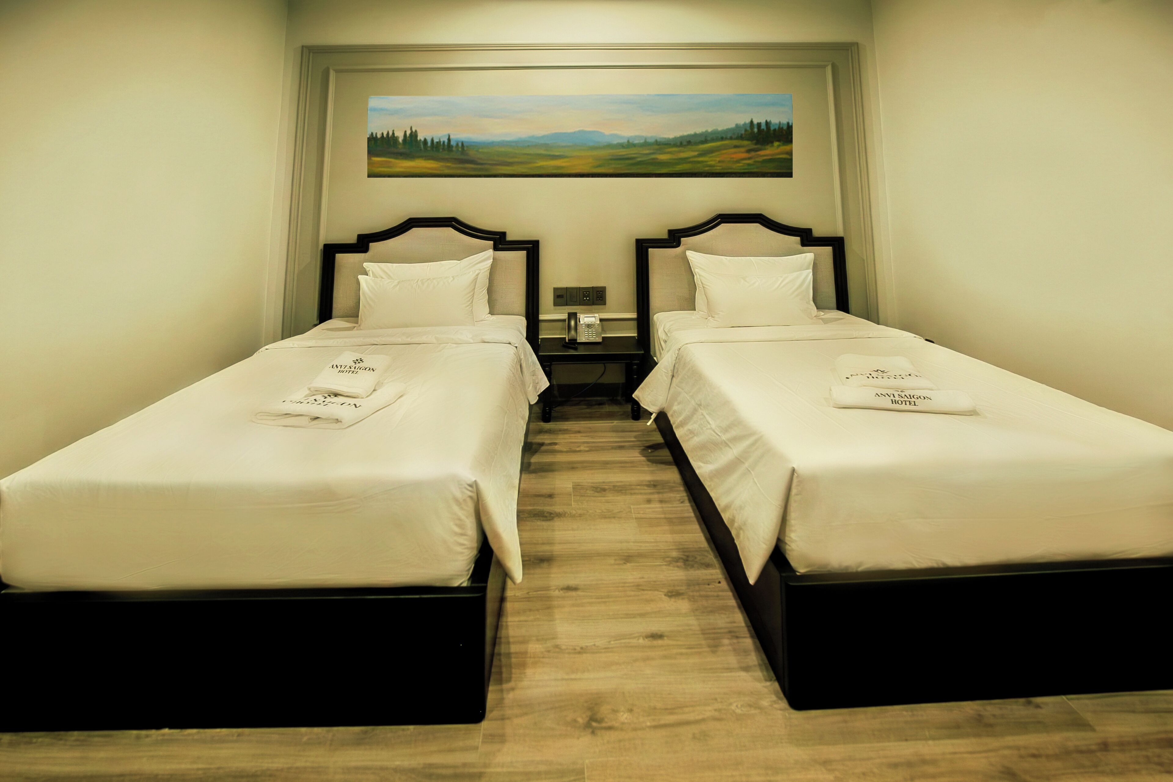 A hotel room with two single beds, a nightstand with a telephone, and a painting of a landscape on the wall.