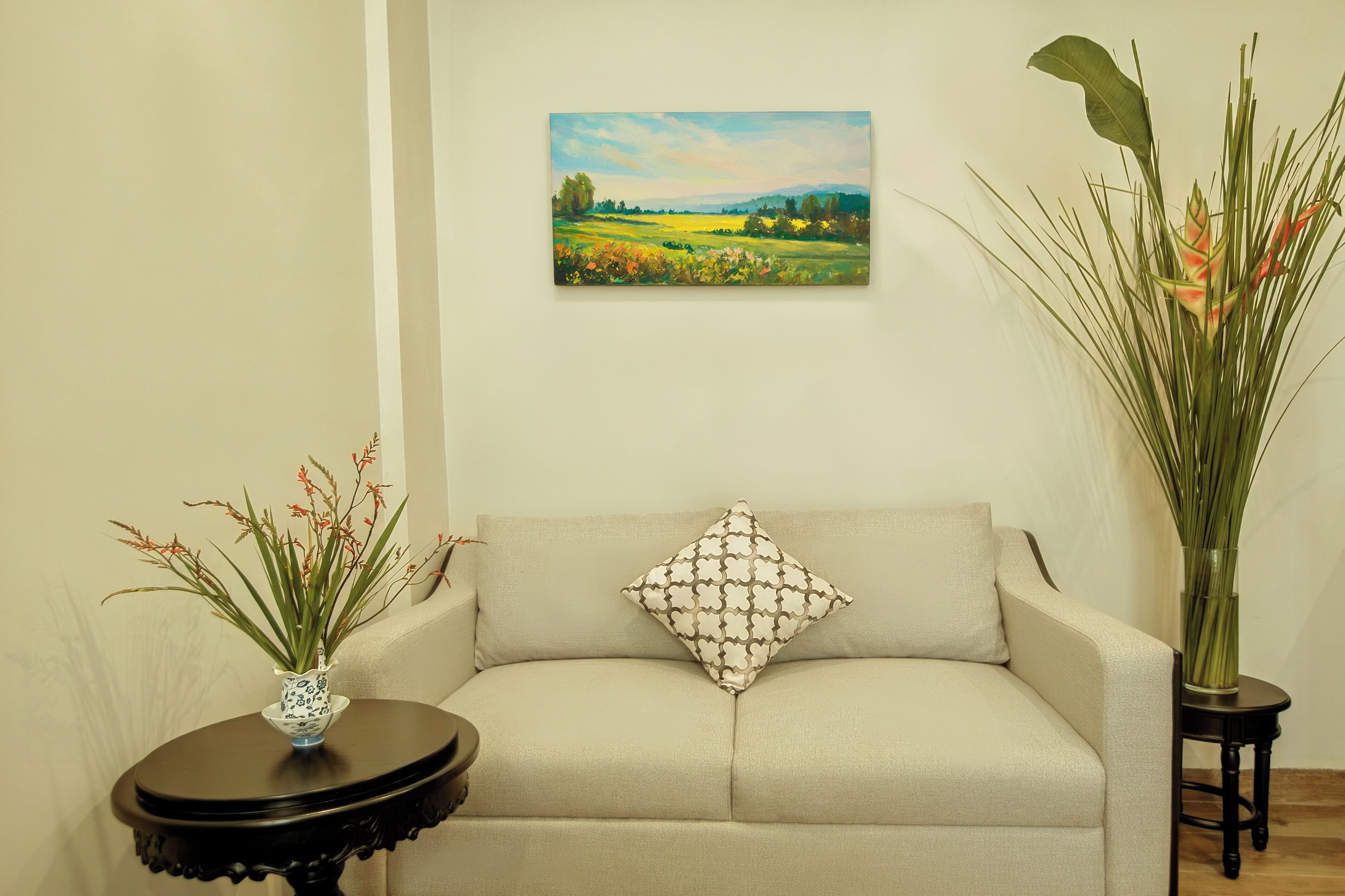 Un soggiorno con un divano color beige, un cuscino decorato, un quadro raffigurante un paesaggio e due vasi con piante.