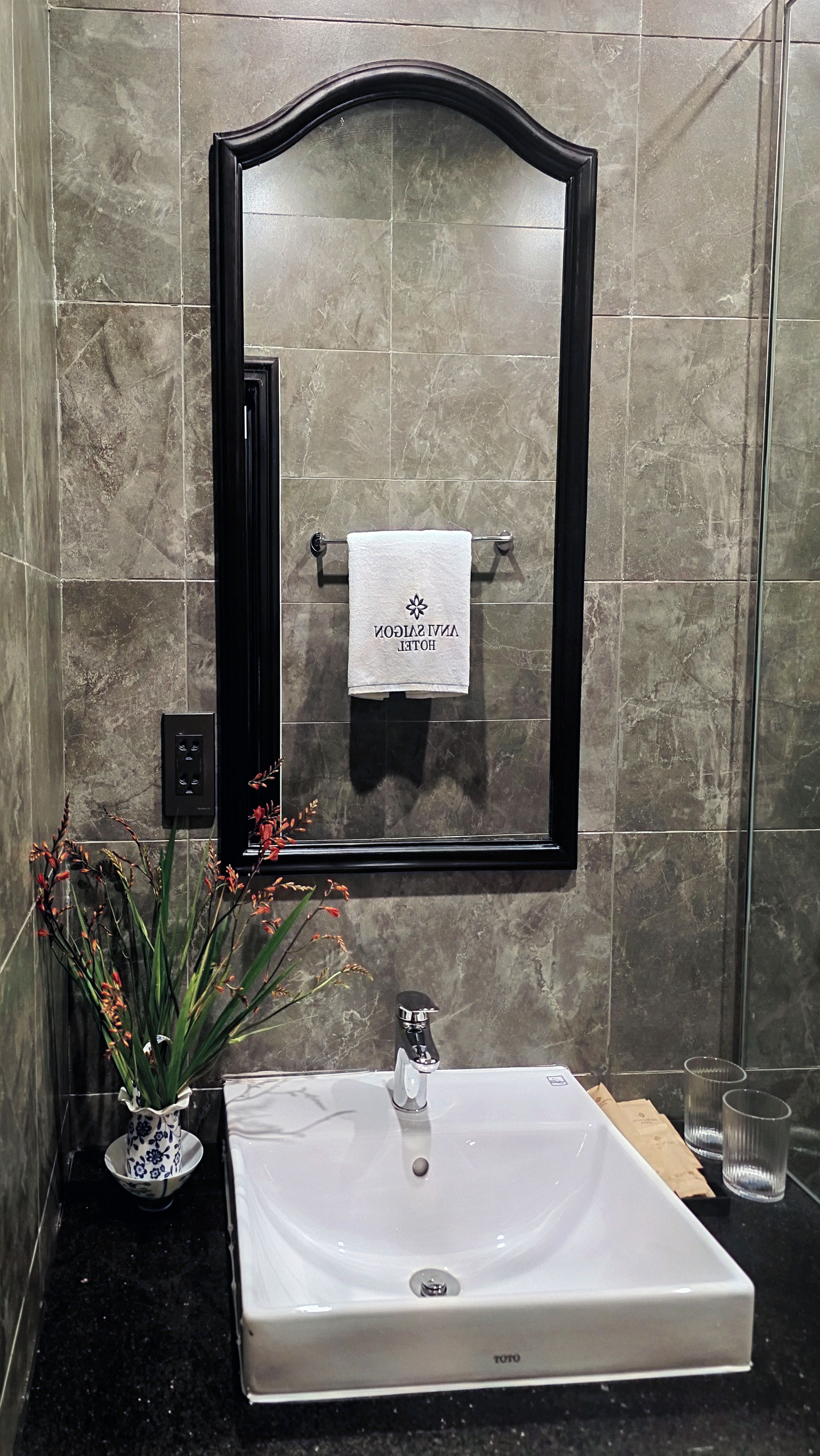 Casa de banho com lavatório branco, espelho com toalha e um vaso com flores.
