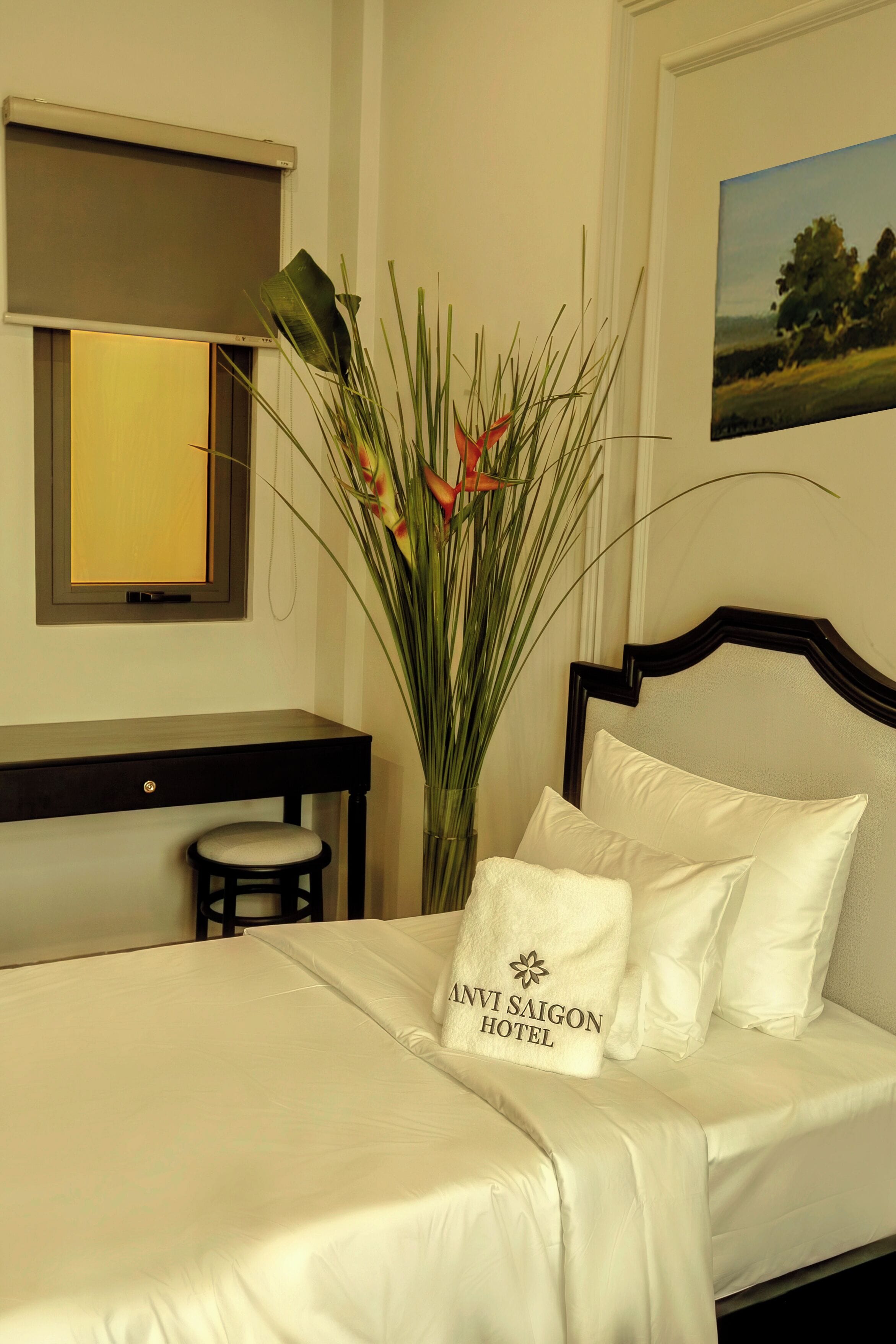 Habitación de hotel con cama, escritorio, silla, una planta grande en maceta y un cuadro en la pared.