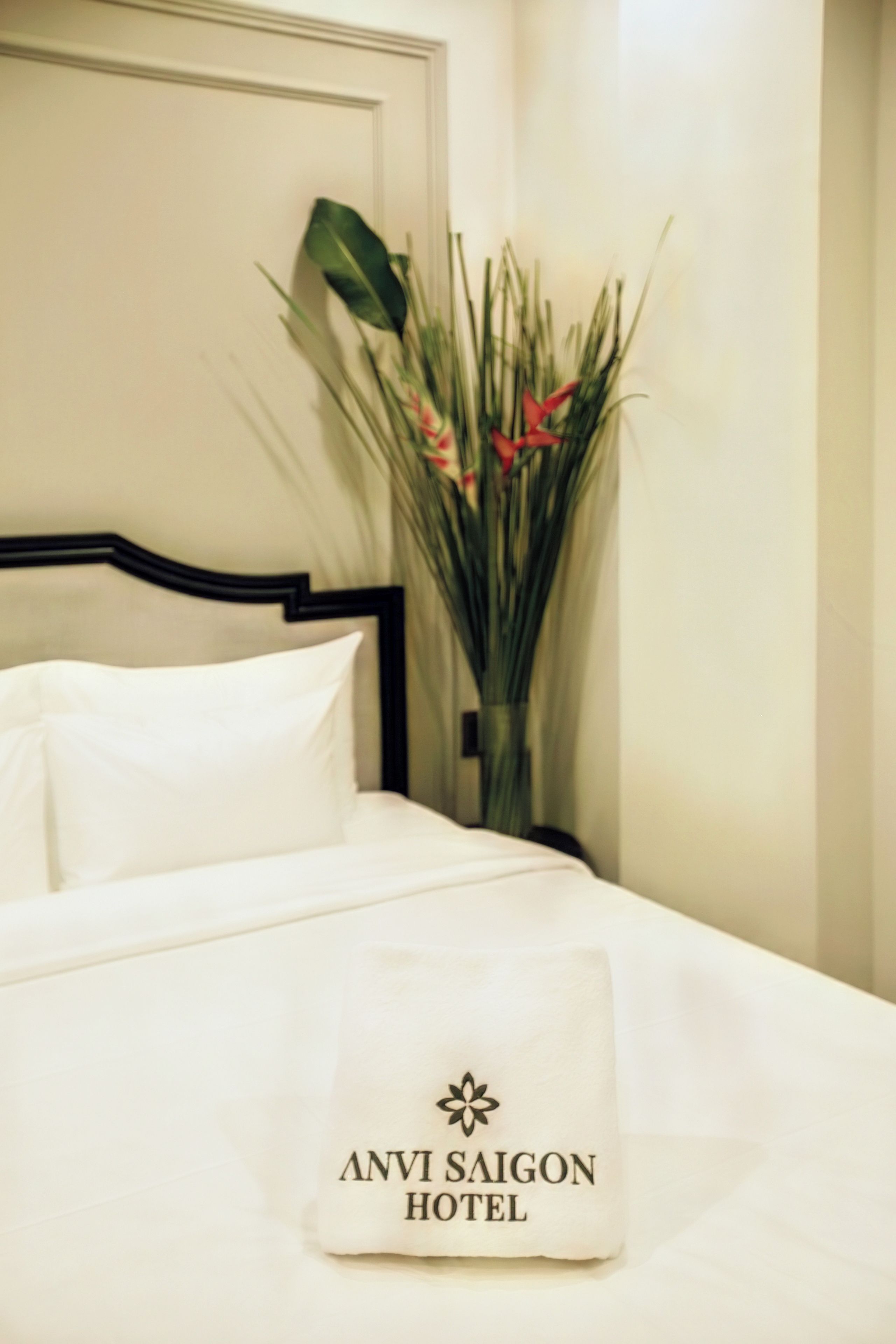 A neatly made bed with white linens and a pillow displaying 'ANVI SAIGON HOTEL' branding.