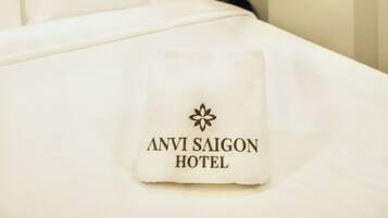 A neatly made bed with white linens and a pillow displaying 'ANVI SAIGON HOTEL' branding.