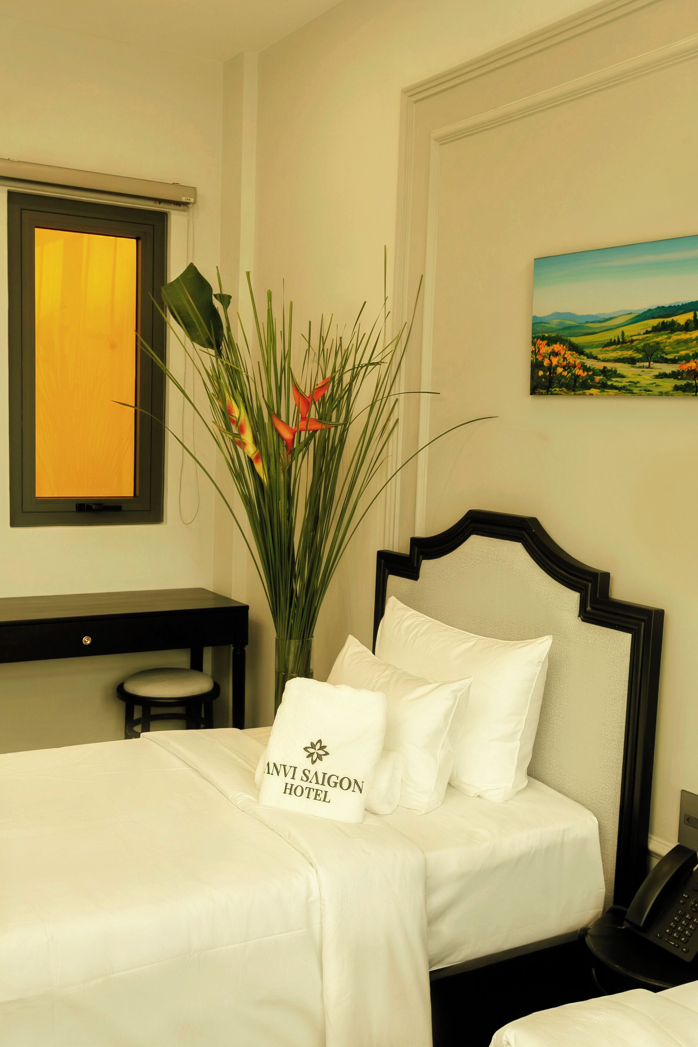 A hotel room with a bed, a desk, a vase with plants, and a painting on the wall.