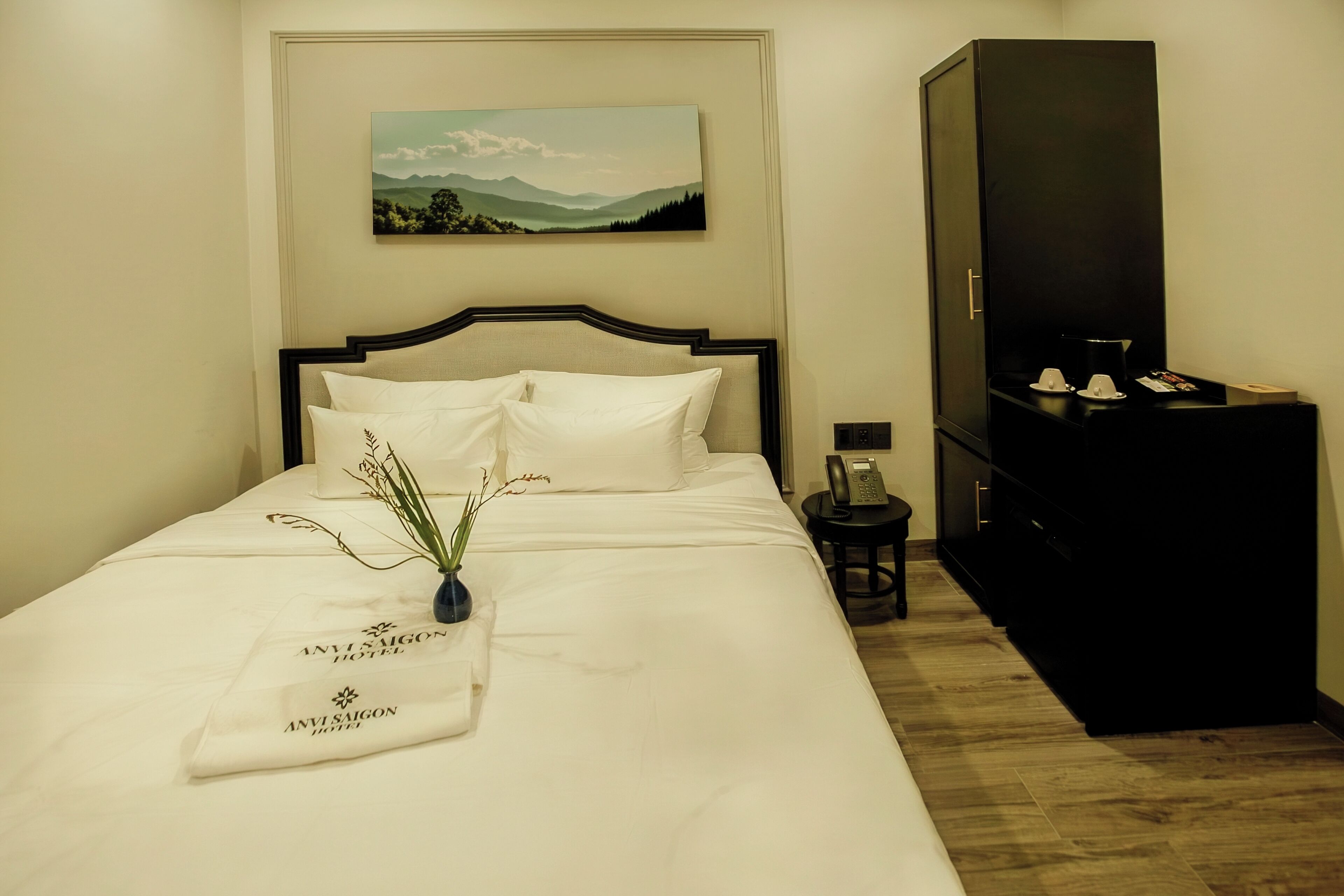 A neatly made bed with white linens, a small bedside table with a lamp, a painting of a landscape on the wall, and a black cabinet.