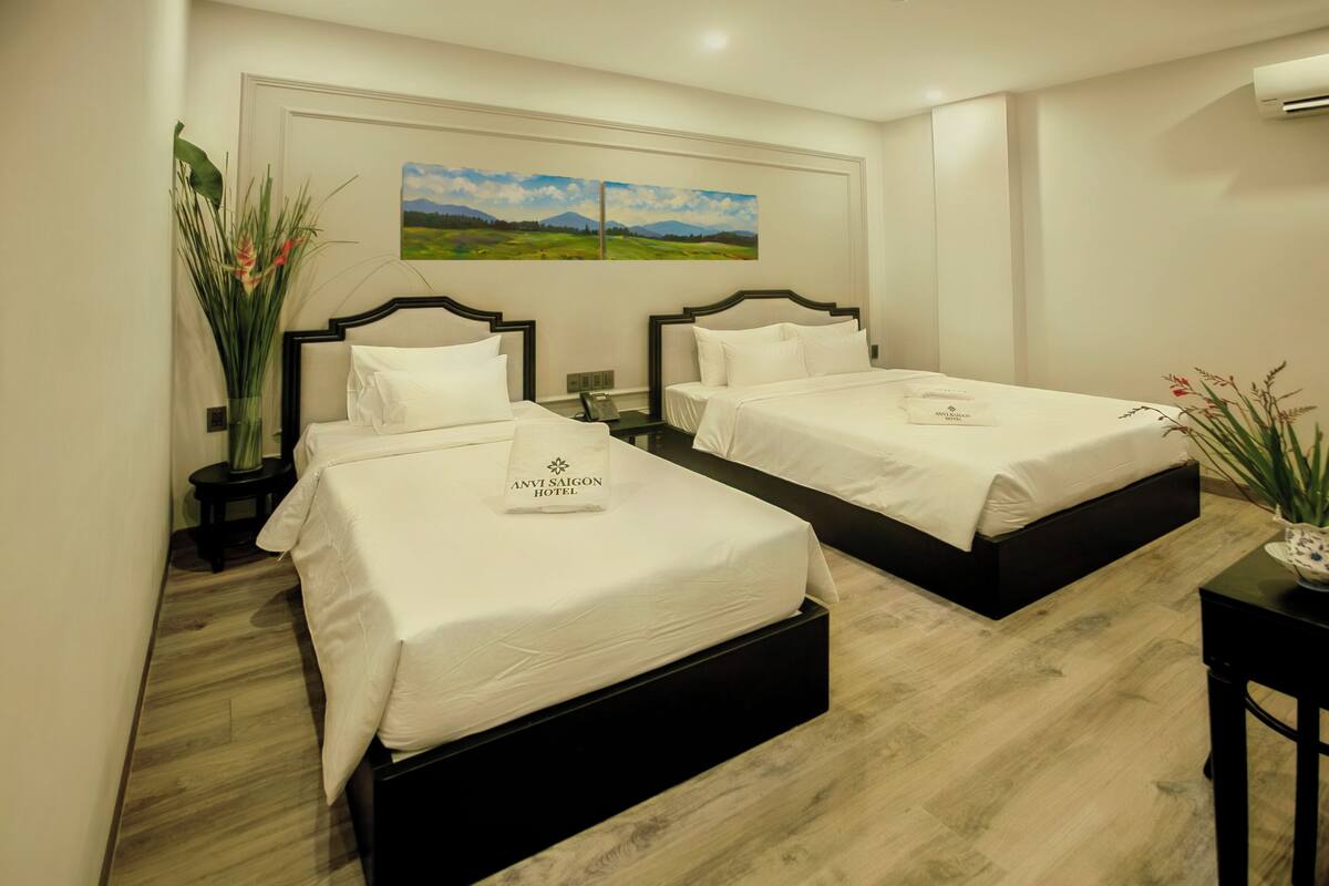 A hotel room with two beds, a nightstand with a phone, a vase with flowers, and a painting on the wall.