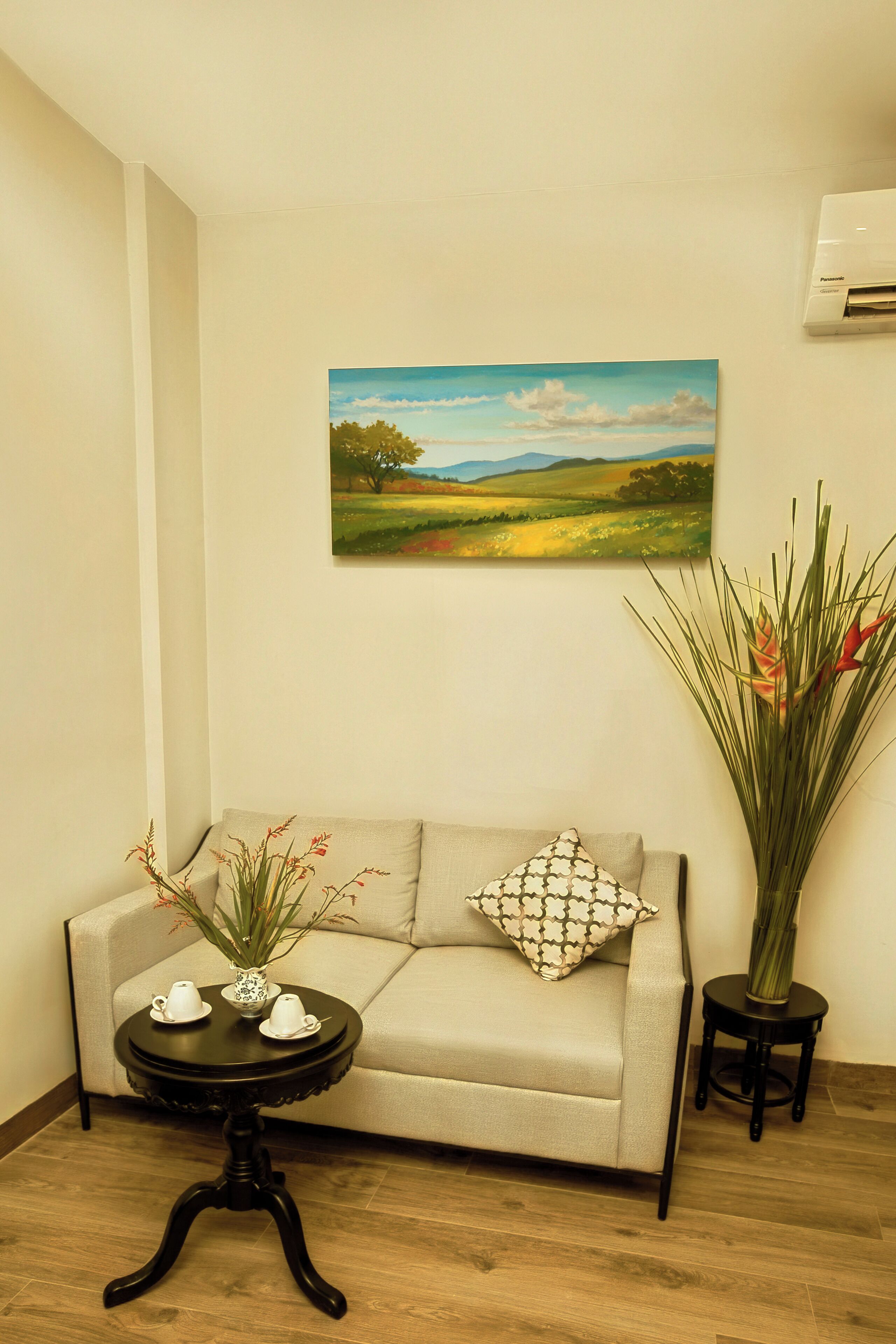 Una habitación con un sofá color crema, una mesita redonda con un florero y tazas de té, y una planta alta sobre una mesita auxiliar.