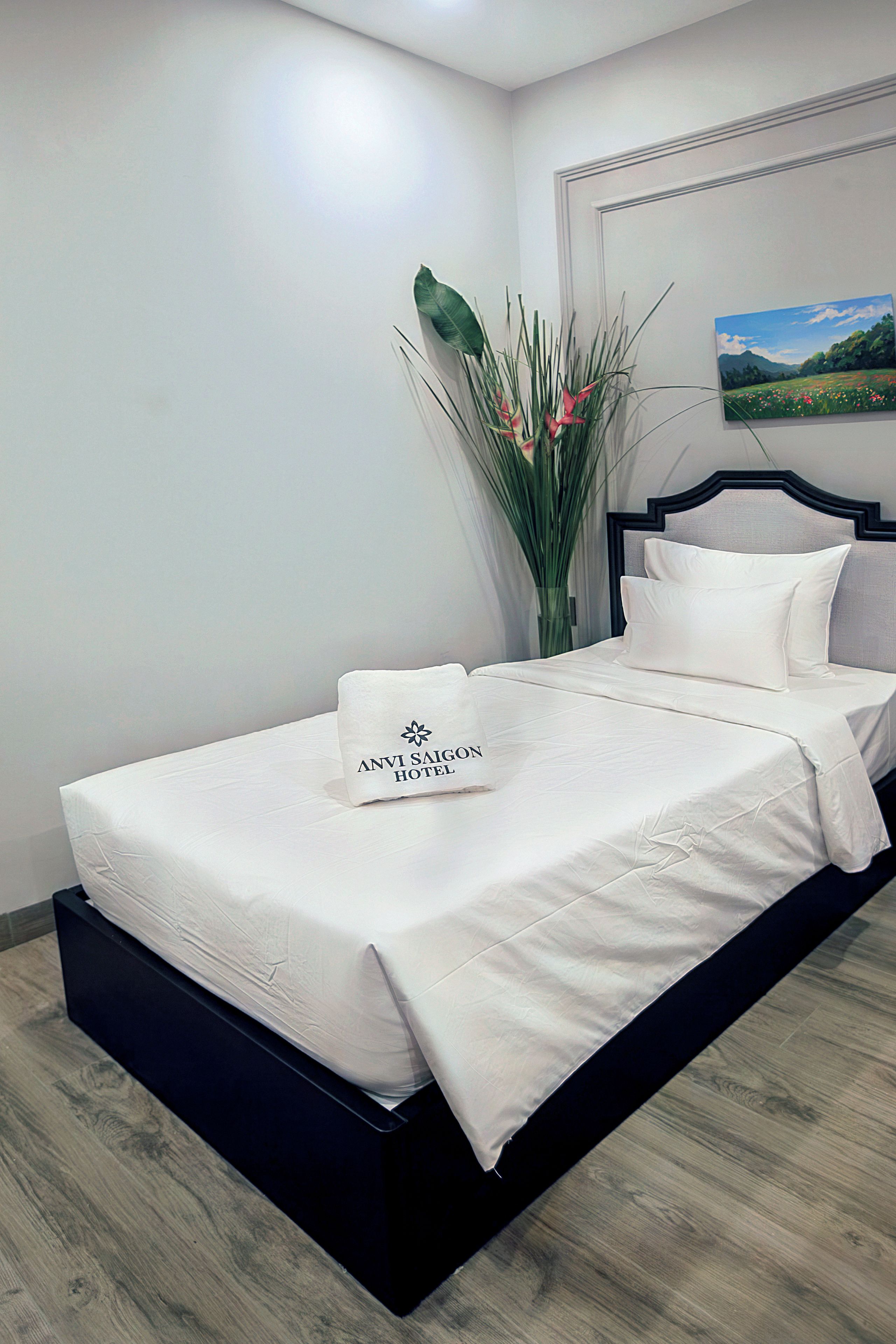 Ein ordentlich bezogenes Bett mit einem Kopfkissen, auf dem 'ANVI SAIGON HOTEL' steht, und ein Landschaftsbild an der Wand.