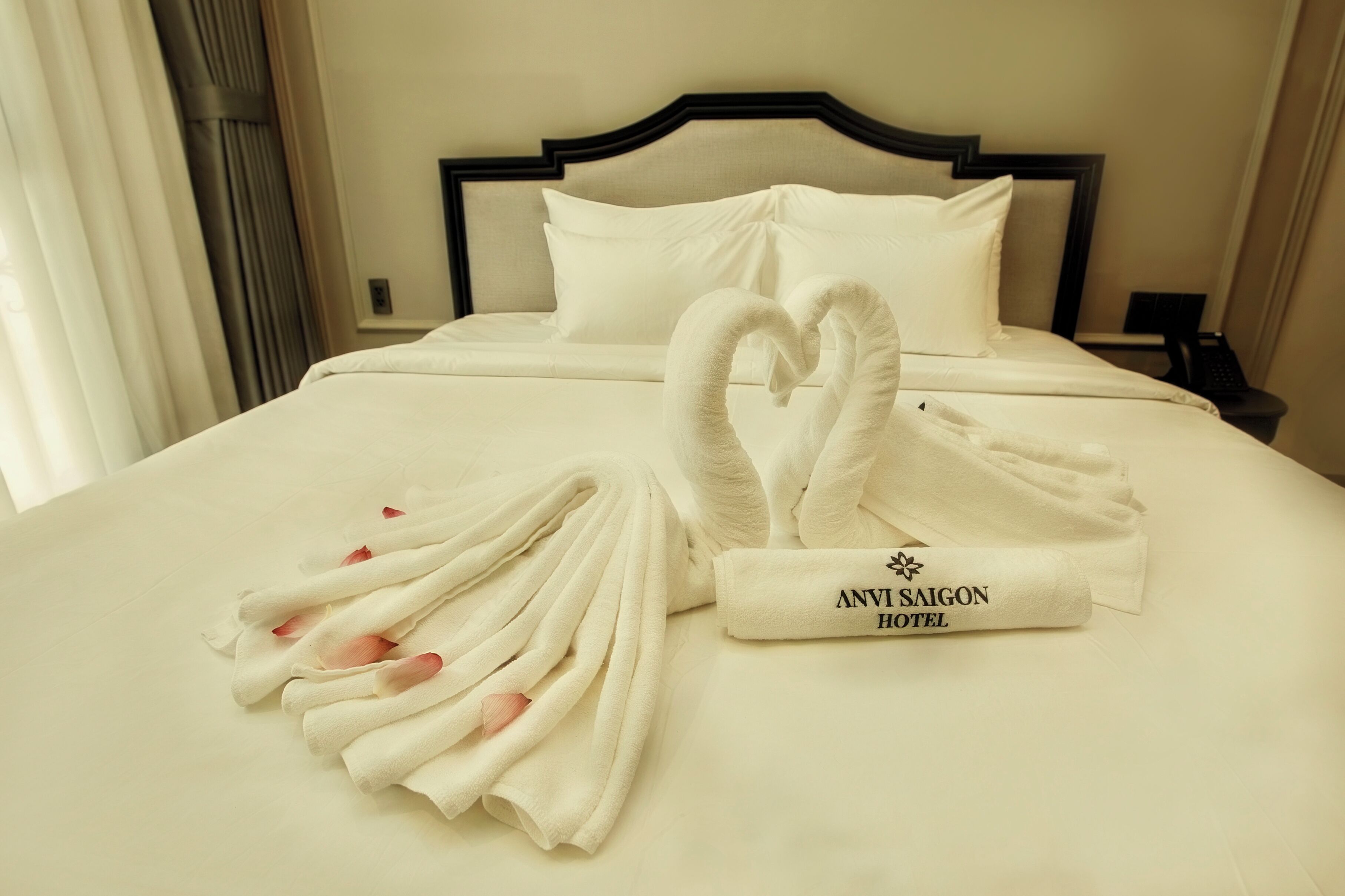 A neatly made bed with a heart-shaped towel arrangement and a rolled towel with 'ANVI SAIGON HOTEL' text.